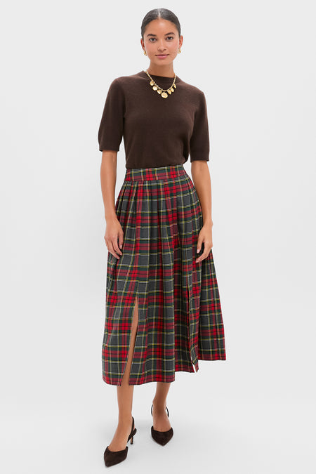 Woman in a brown short-sleeve top, Hunter Bell Bemelmans Plaid skirt with front slit, brown heels, and gold statement necklace stands against a plain light background—a perfect holiday party look.