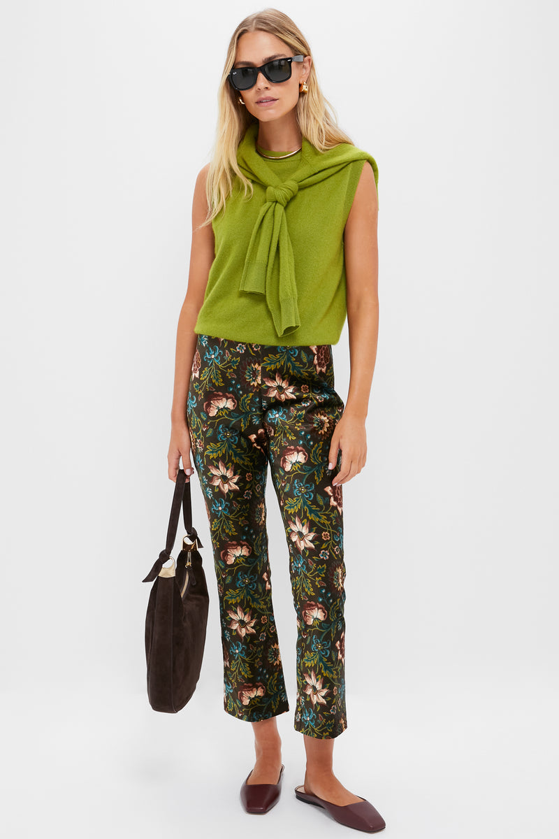 A woman stands against a white background wearing a green sleeveless Tuckernuck sweater with matching scarf, high-rise floral print pants, brown bag, brown flats, and sunglasses.