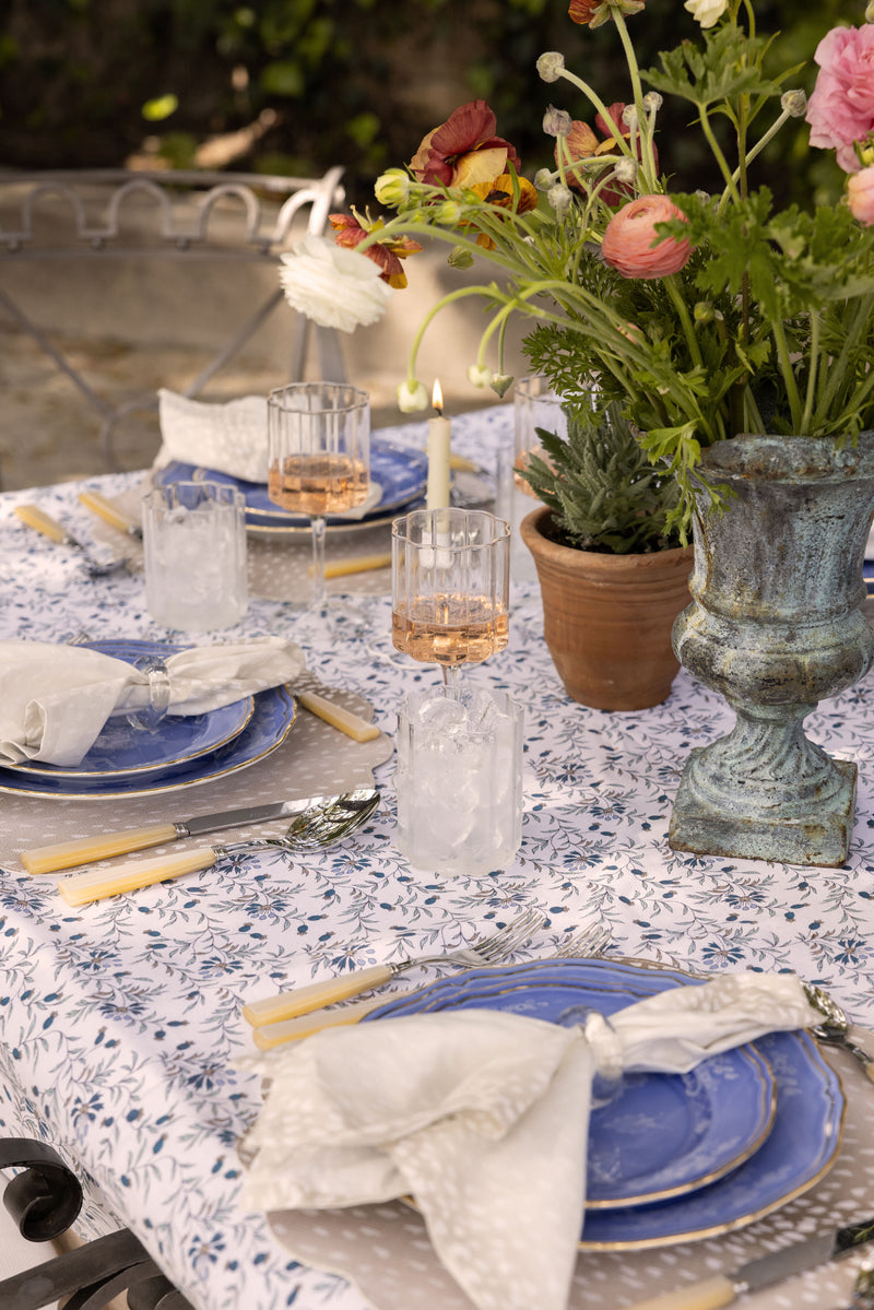 A table set for a meal with blue plates, Tuckernuck Home Antelope Printed Napkins, glasses with ice, a lit candle, and a floral centerpiece on a patterned tablecloth.