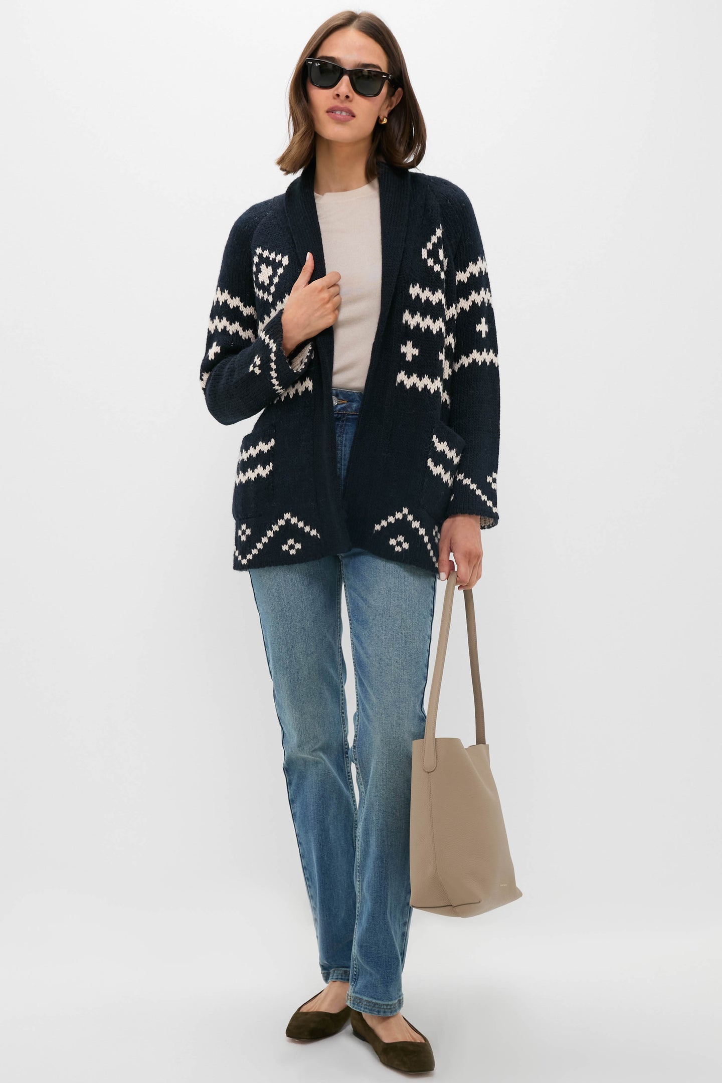 Woman wearing sunglasses, a navy patterned cardigan, light beige top, Vanessa Bruno straight-cut jeans, brown flats, and a beige tote bag stands against a plain white background.