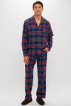 A man wears a Petite Plume classic holiday flannel pajama set in blue, red, and green plaid with black slippers, posed against a plain white background.
