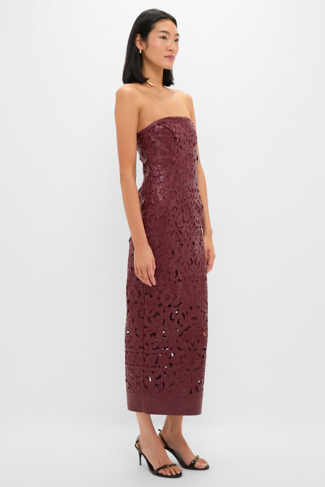 A woman wears a Cult Gaia strapless burgundy patterned midi dress with cutout details and faux leather lace accents, paired with black heeled sandals—perfect for chic evening events on a plain white background.