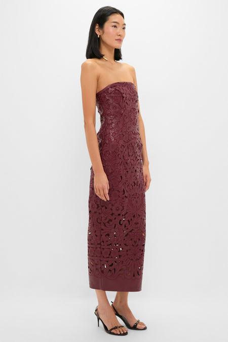 A woman wears a Cult Gaia strapless burgundy patterned midi dress with cutout details and faux leather lace accents, paired with black heeled sandals—perfect for chic evening events on a plain white background.