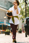 Woman walking with baguettes in a city in a white button down, green sweater, and burgundy pants. 
