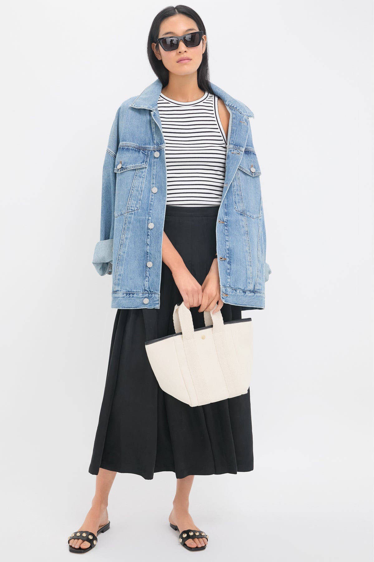 A woman wears a black pleated maxi skirt, styled with a denim jacket, striped tank top, black sunglasses, a white tote bag, and sandals.