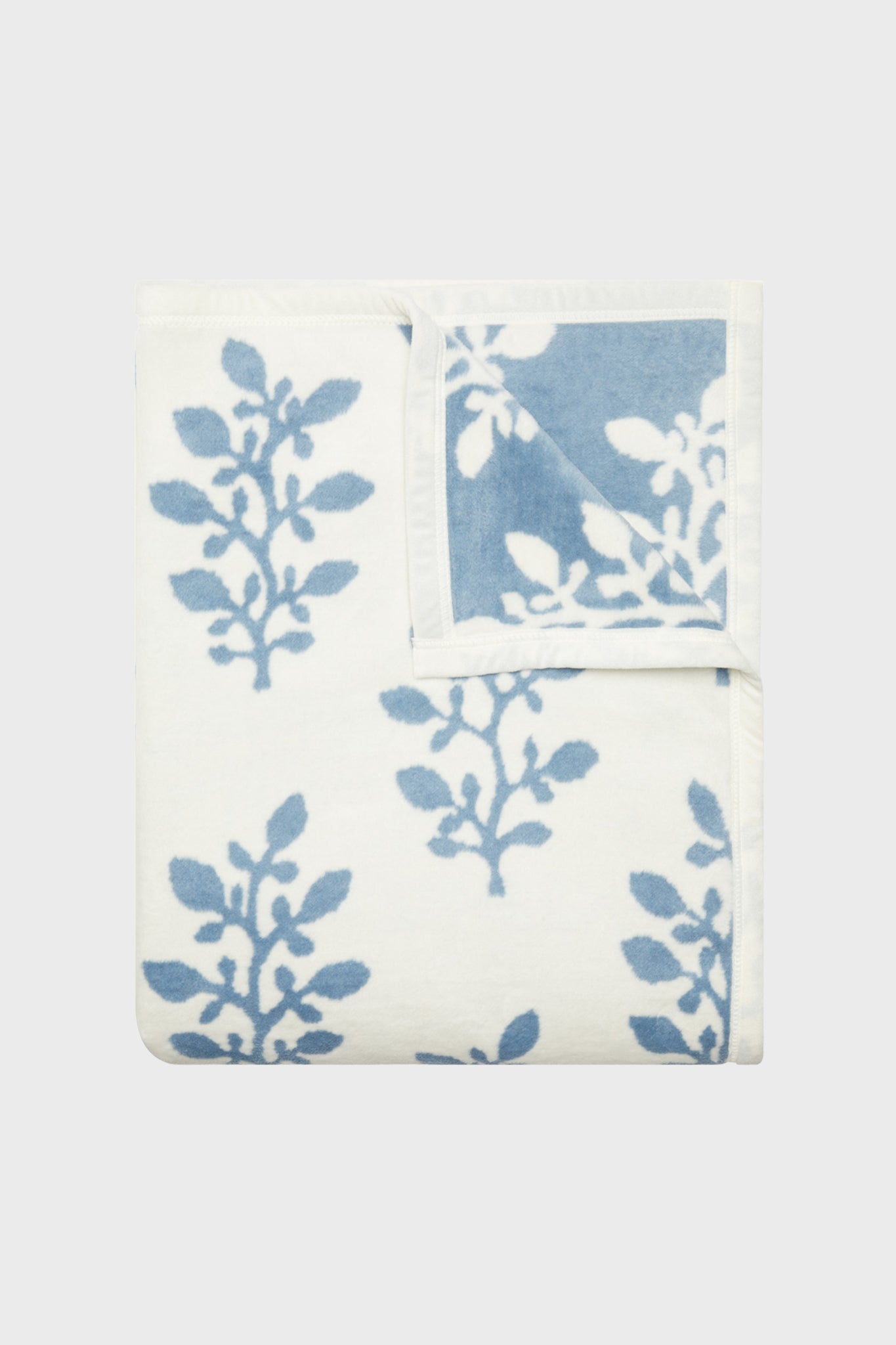 A folded ChappyWrap Garden Gate Blanket displays its blue and white leaf pattern, with a flipped corner showing the reverse. This stylish throw from ChappyWrap is machine washable and versatile for any setting.