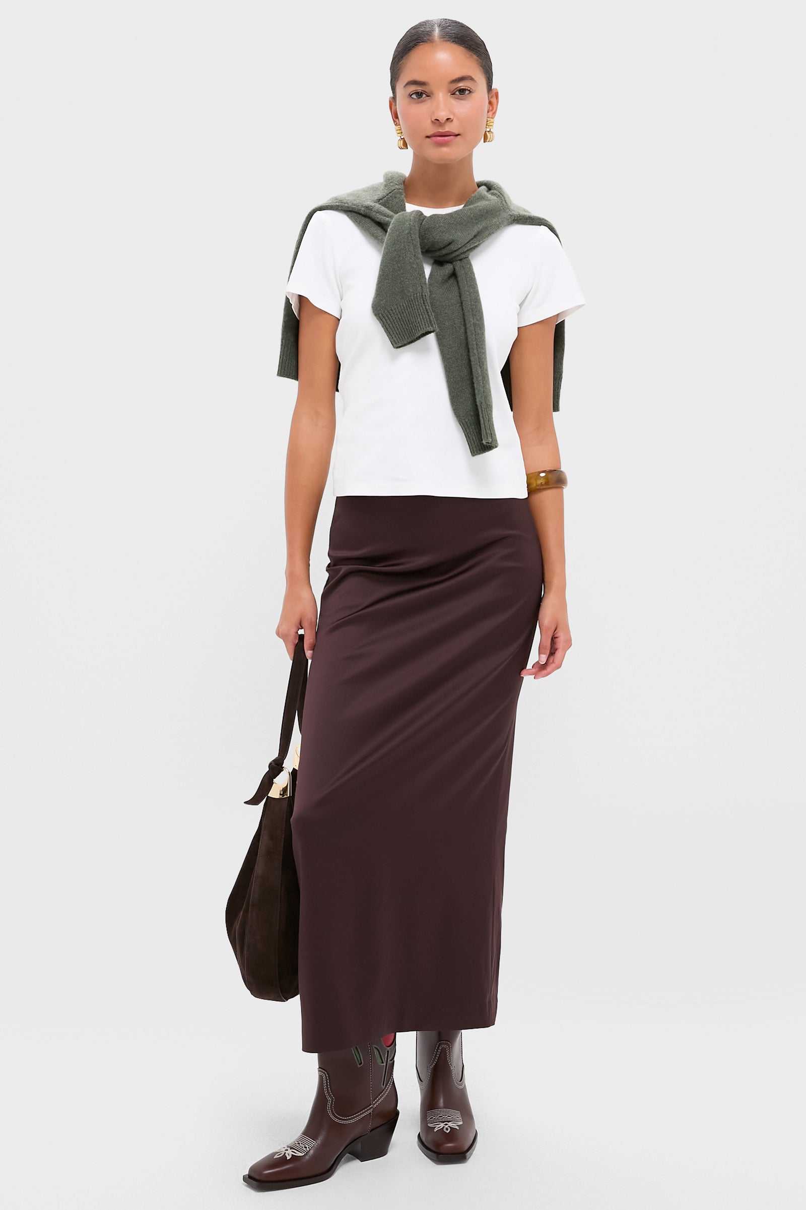 A woman wears a white t-shirt, olive green sweater over her shoulders, long brown skirt, Loeffler Randall boots, and carries a brown bag.