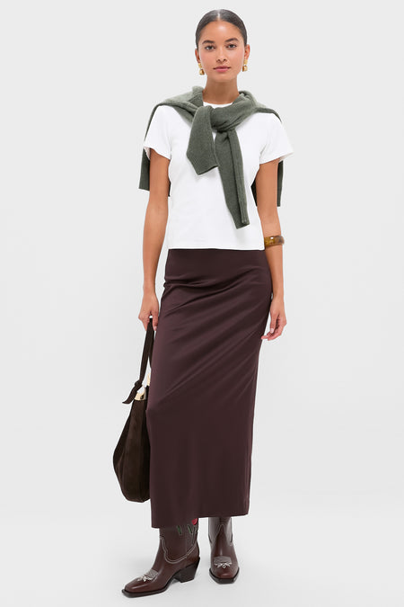 A woman wears a white t-shirt, olive green sweater over her shoulders, long brown skirt, Loeffler Randall boots, and carries a brown bag.