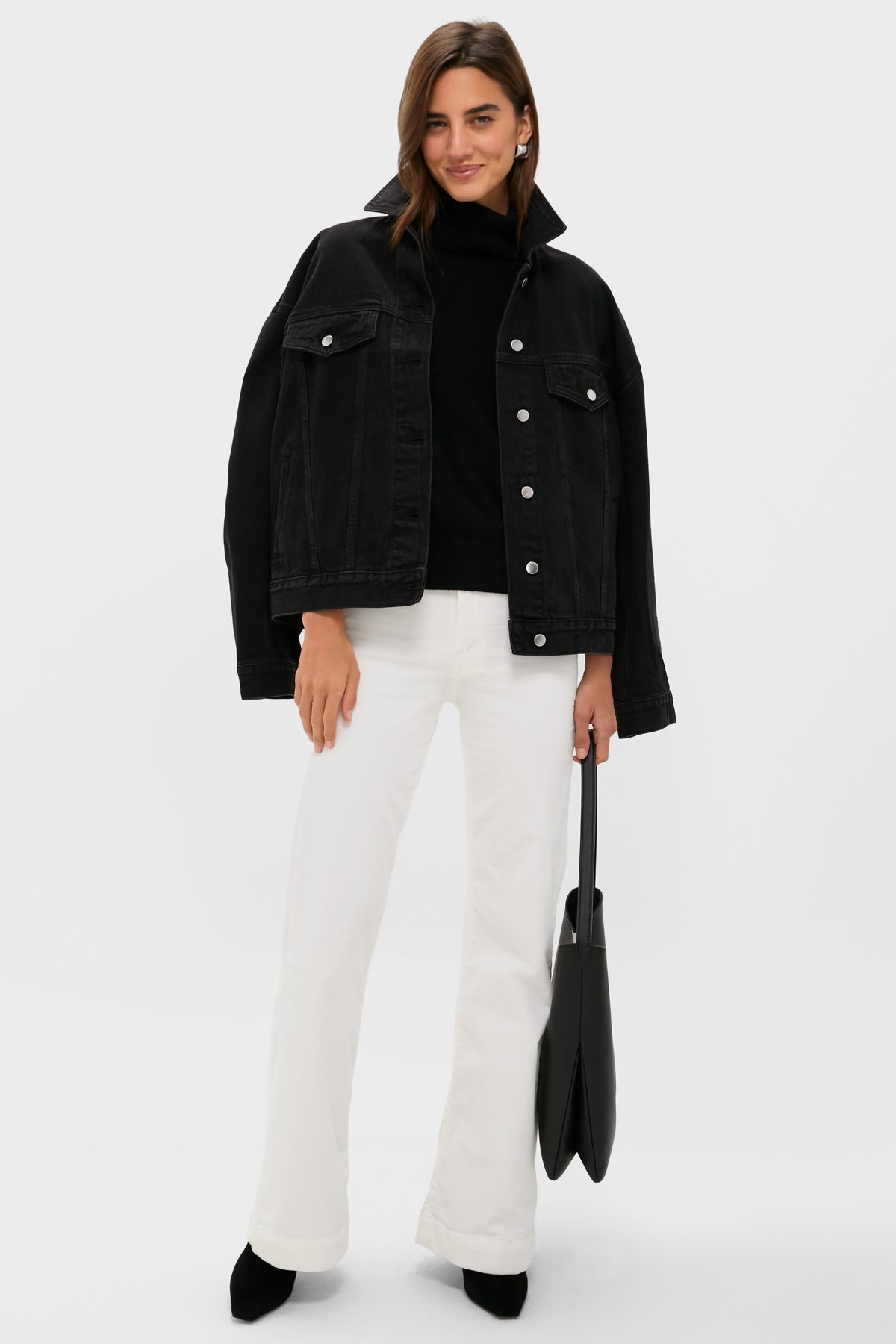 A woman stands against a plain background wearing a black denim jacket, black top, ASKK NY high-rise wide leg pants, black shoes, and holding a black tote bag from ASKK NY.