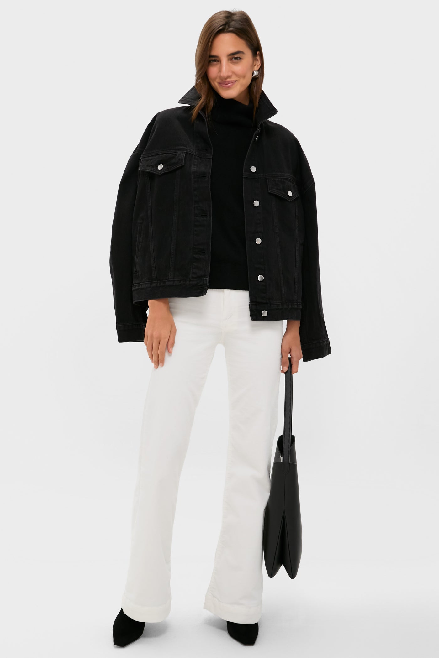 A woman stands against a plain background wearing a black denim jacket, black top, ASKK NY high-rise wide leg pants, black shoes, and holding a black tote bag from ASKK NY.