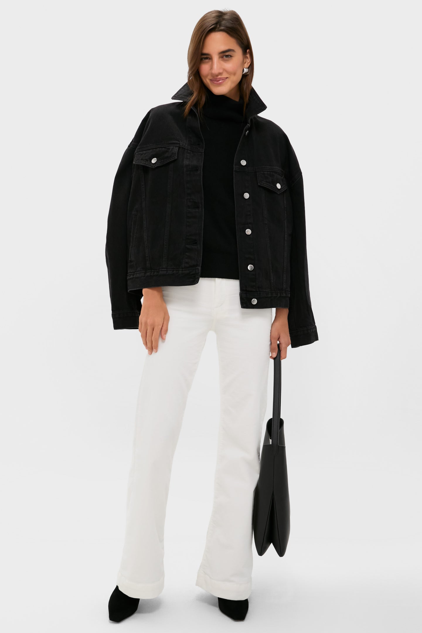 A woman stands against a plain background wearing a black denim jacket, black top, ASKK NY high-rise wide leg pants, black shoes, and holding a black tote bag from ASKK NY.