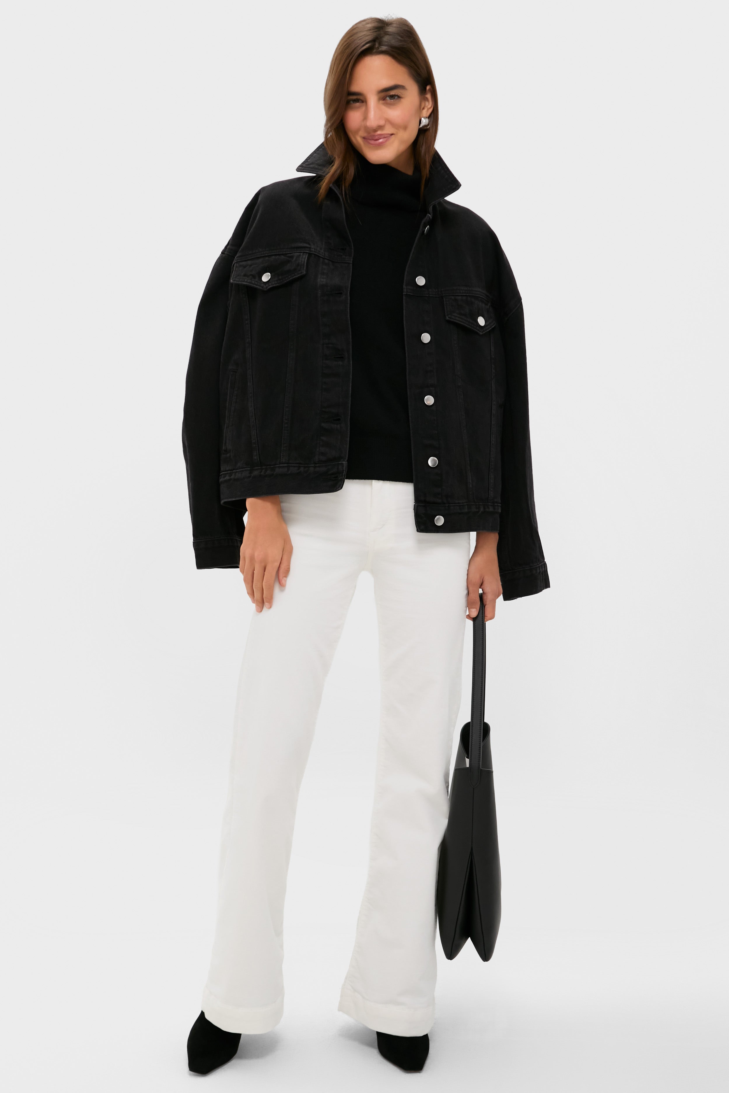 A woman stands against a plain background wearing a black denim jacket, black top, ASKK NY high-rise wide leg pants, black shoes, and holding a black tote bag from ASKK NY.