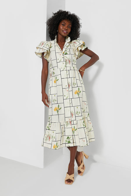 A woman wears a white, grid-patterned Tuckernuck cotton midi dress with floral prints and ruffle sleeves, paired with beige heeled sandals.