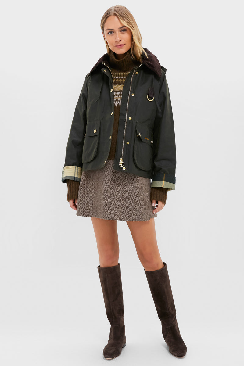 A woman in a Barbour Reighton Spey Waxed Jacket, brown sweater, patterned mini skirt, and knee-high brown boots faces forward against a plain light background.