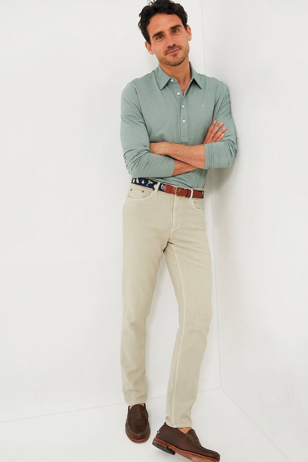 Man with arms crossed and a neutral expression, wearing a light green Criquet performance long sleeve polo, beige pants, brown loafers, and a brown belt.