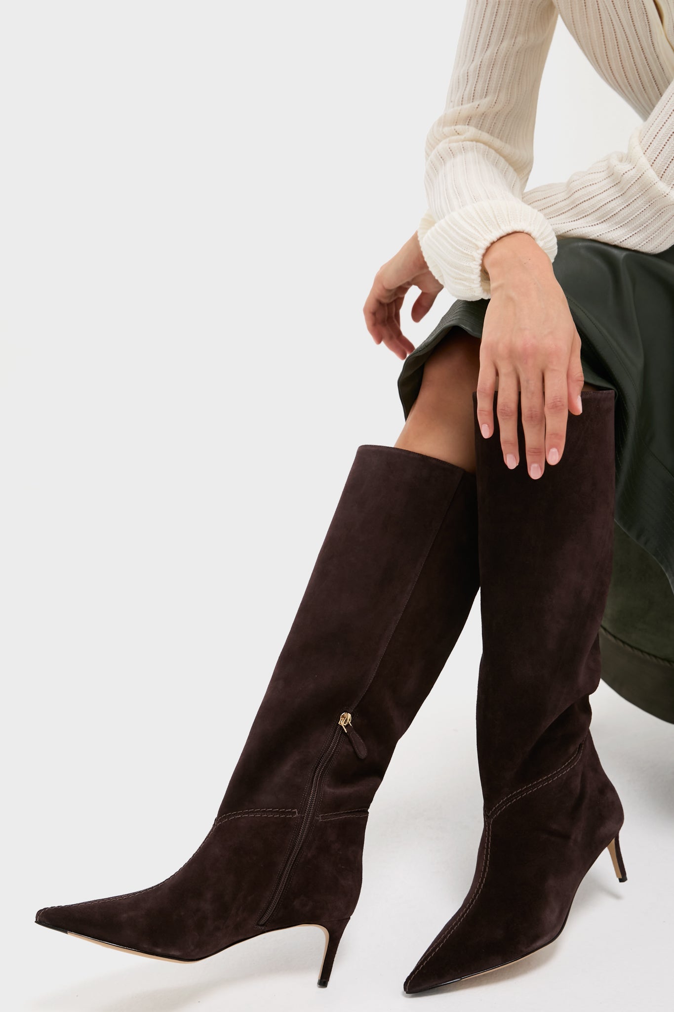 A person sits wearing Alexandre Birman dark brown suede knee-high boots with pointed toes and kitten heels, styled with a long dark skirt and a cream ribbed top.