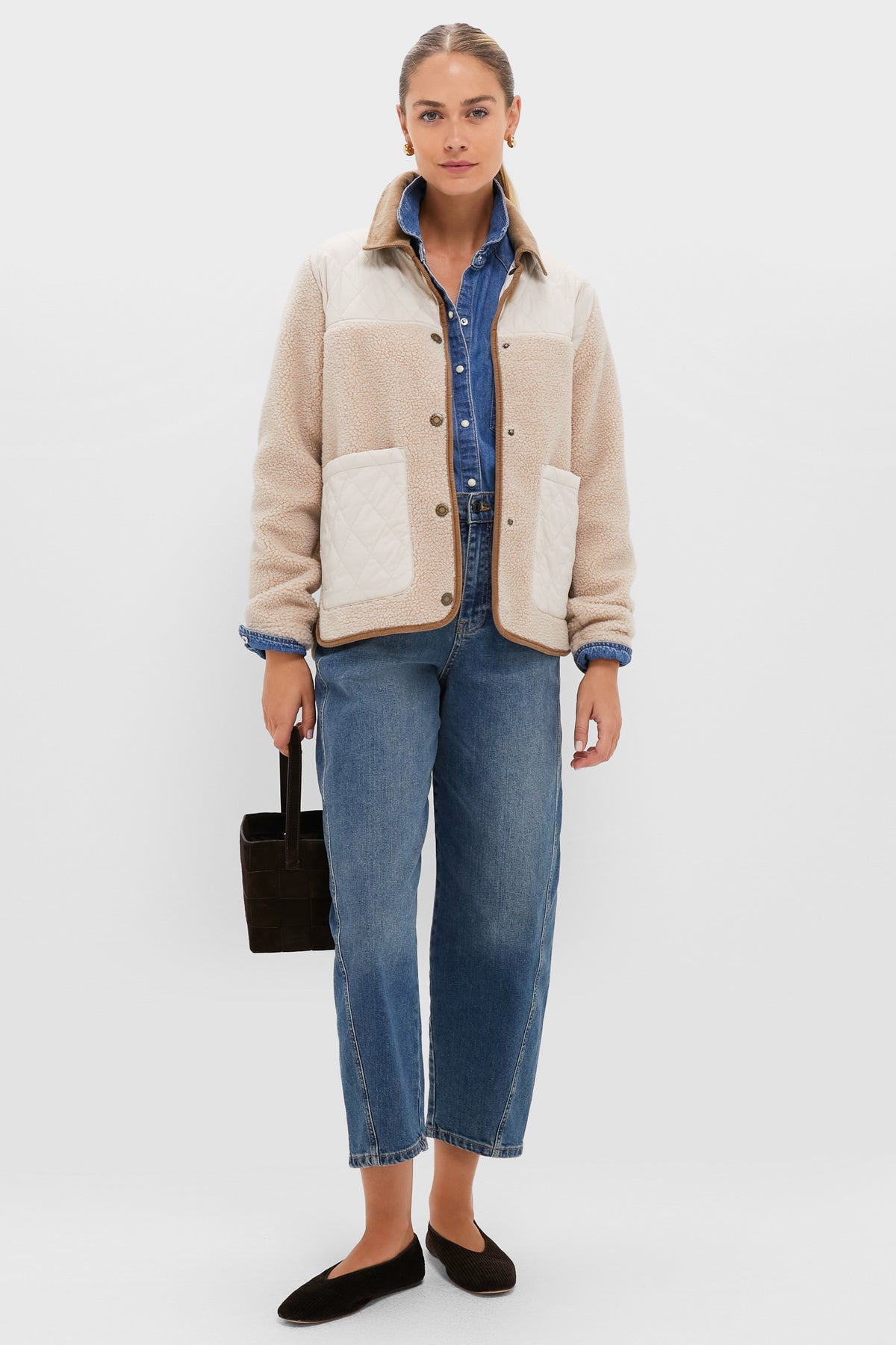 A woman wears a beige fleece jacket, blue denim shirt, blue jeans, and black flats, styled with a black handbag.