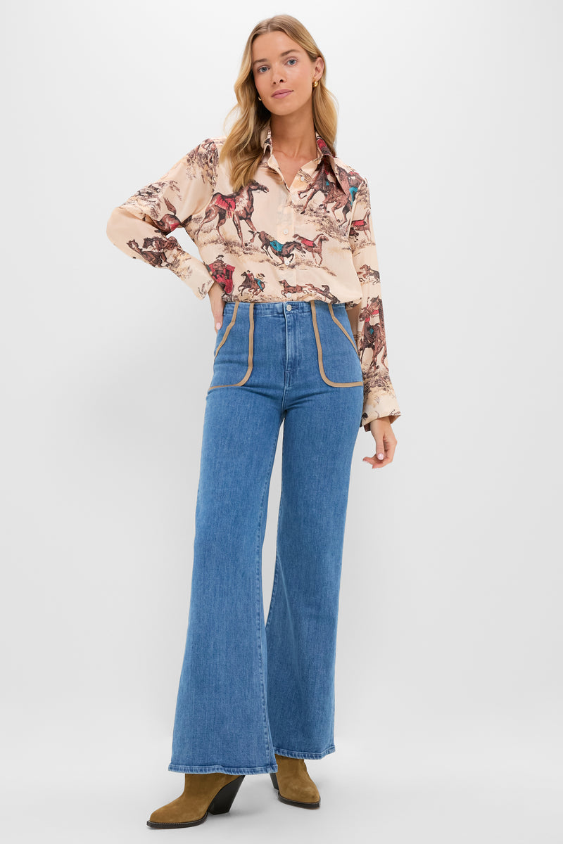 A woman poses against a plain backdrop in LE JEAN's horse-printed blouse, high-rise flare jeans in blue crystal denim, and tan heeled boots. Made in Los Angeles.