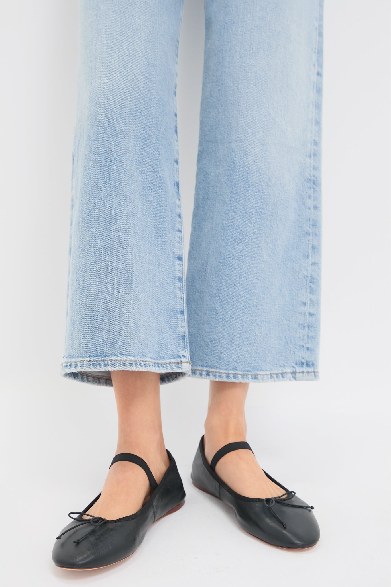 A person stands against a white background wearing light blue wide-leg jeans and Loeffler Randall black Leonie Flat leather ballet flats with a strap.