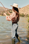 Woman in the water fishing. She's wearing a straw cowboy hat, a plaid shirt, jeans with a leather belt, and rainboots.
