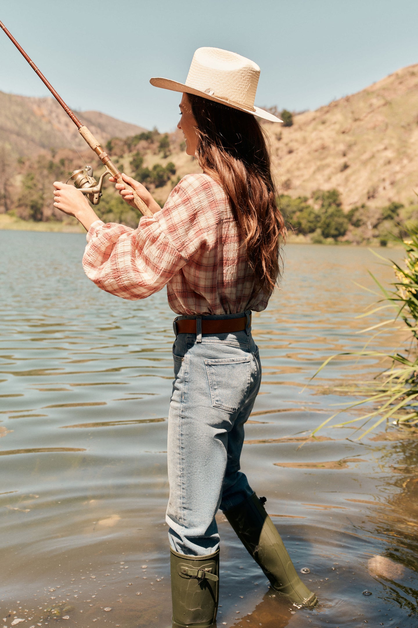 Woman in the water fishing. She's wearing a straw cowboy hat, a plaid shirt, jeans with a leather belt, and rainboots.