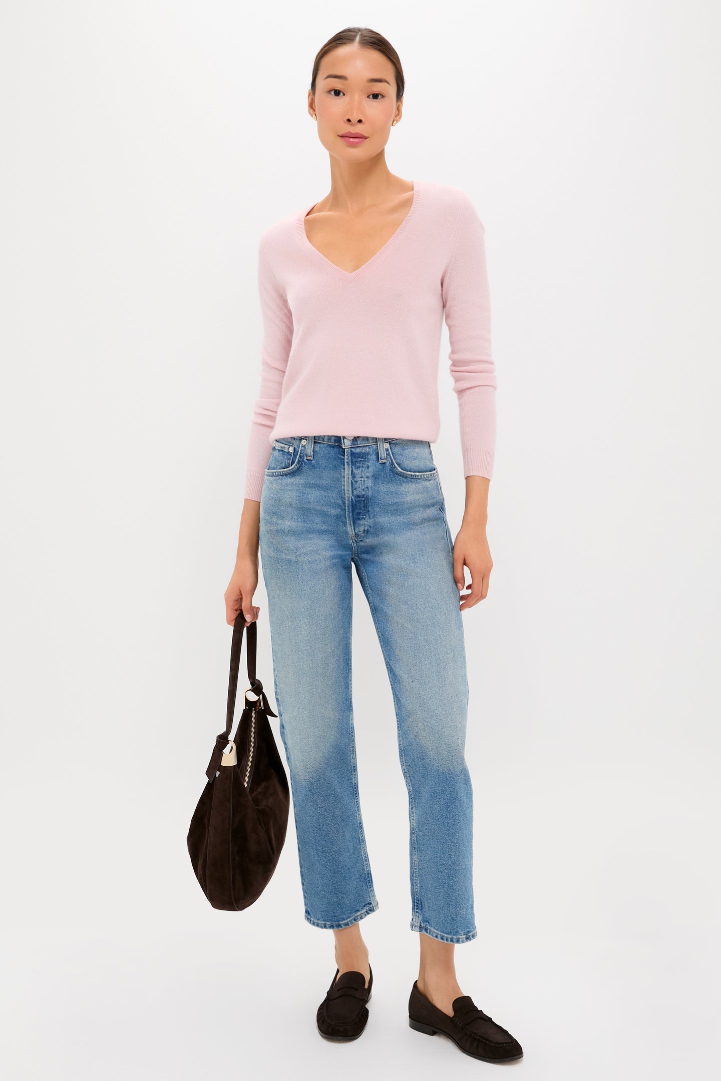 A woman stands against a plain background wearing a Tuckernuck soft pink cashmere V-neck sweater, blue jeans, black loafers, and holding a black handbag—showcasing affordable luxury in everyday style.