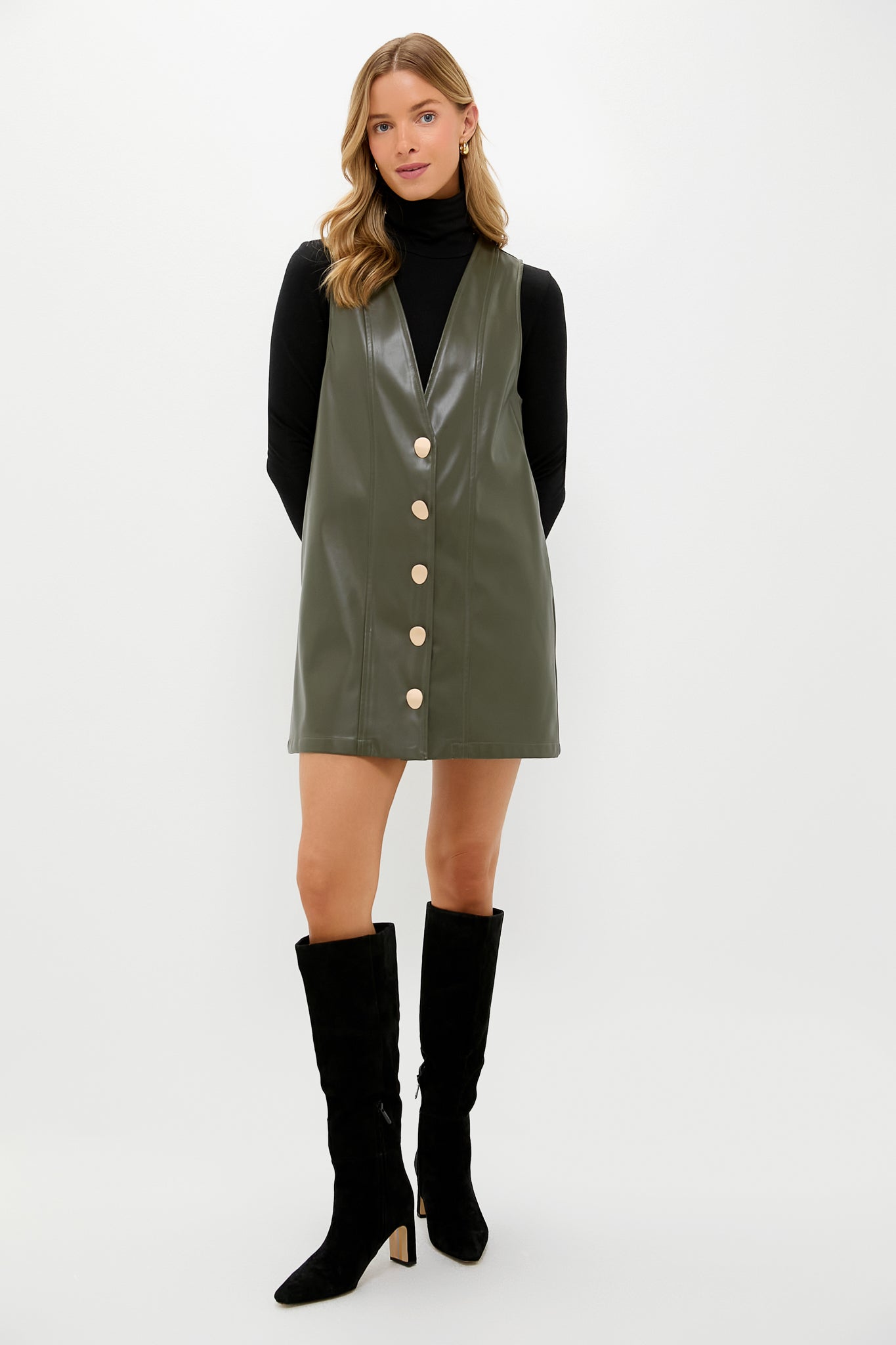 Model in a classic olive faux leather pinafore dress over a black turtleneck, styled with knee-high suede boots, embodying effortless, fun, high-end fashion.