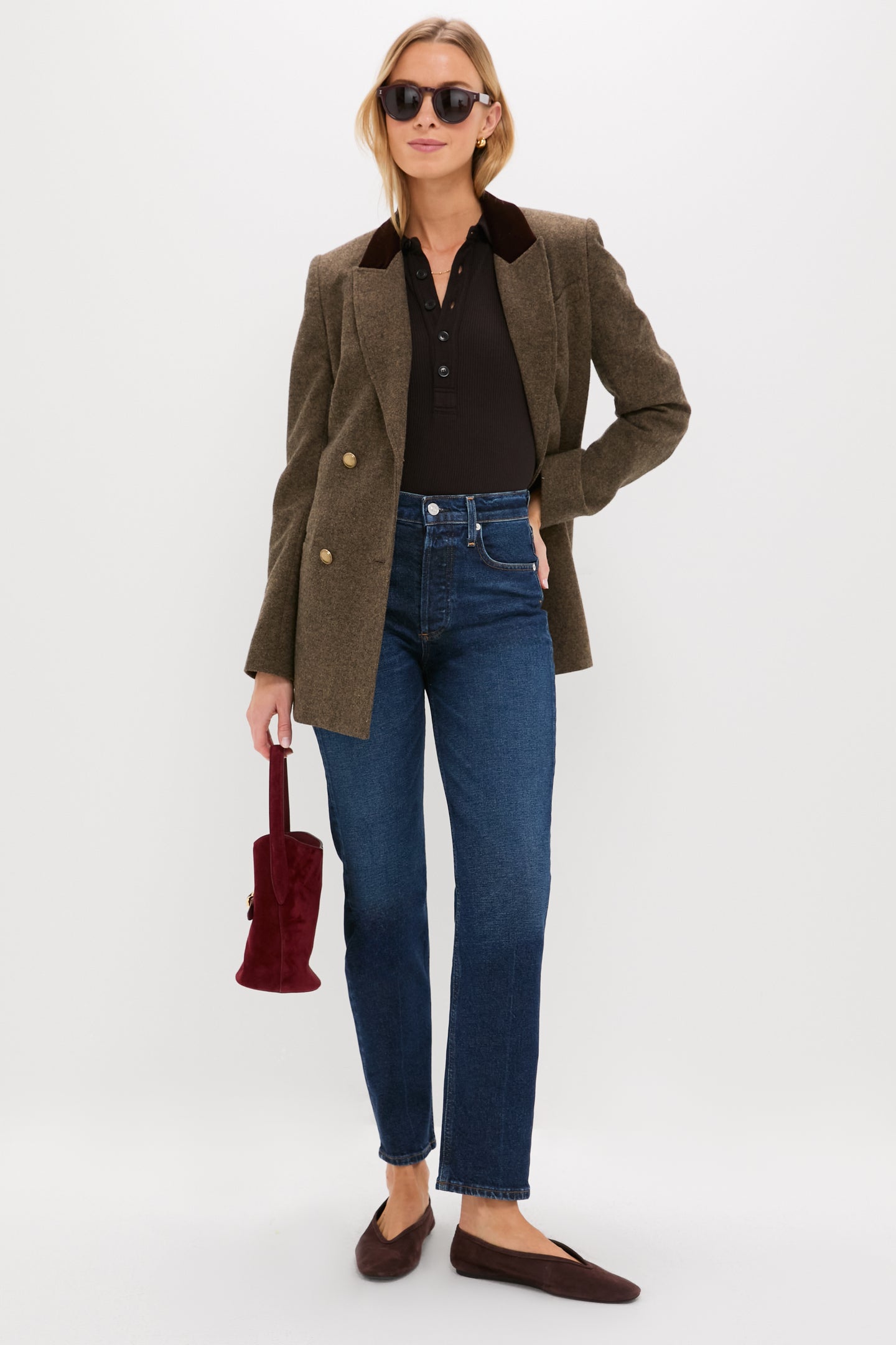 Wearing sunglasses, a brown blazer over a black ribbed cotton long-sleeve polo, blue Citizens of Humanity jeans, and brown flats, a person with a small red handbag stands against a plain white background.
