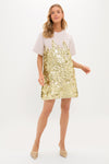 A woman wears the Hunter Bell Aerin Dress, a short-sleeved, light-colored evening shift with gold sequins on the lower half, paired with pointed-toe shoes, standing against a plain white background.