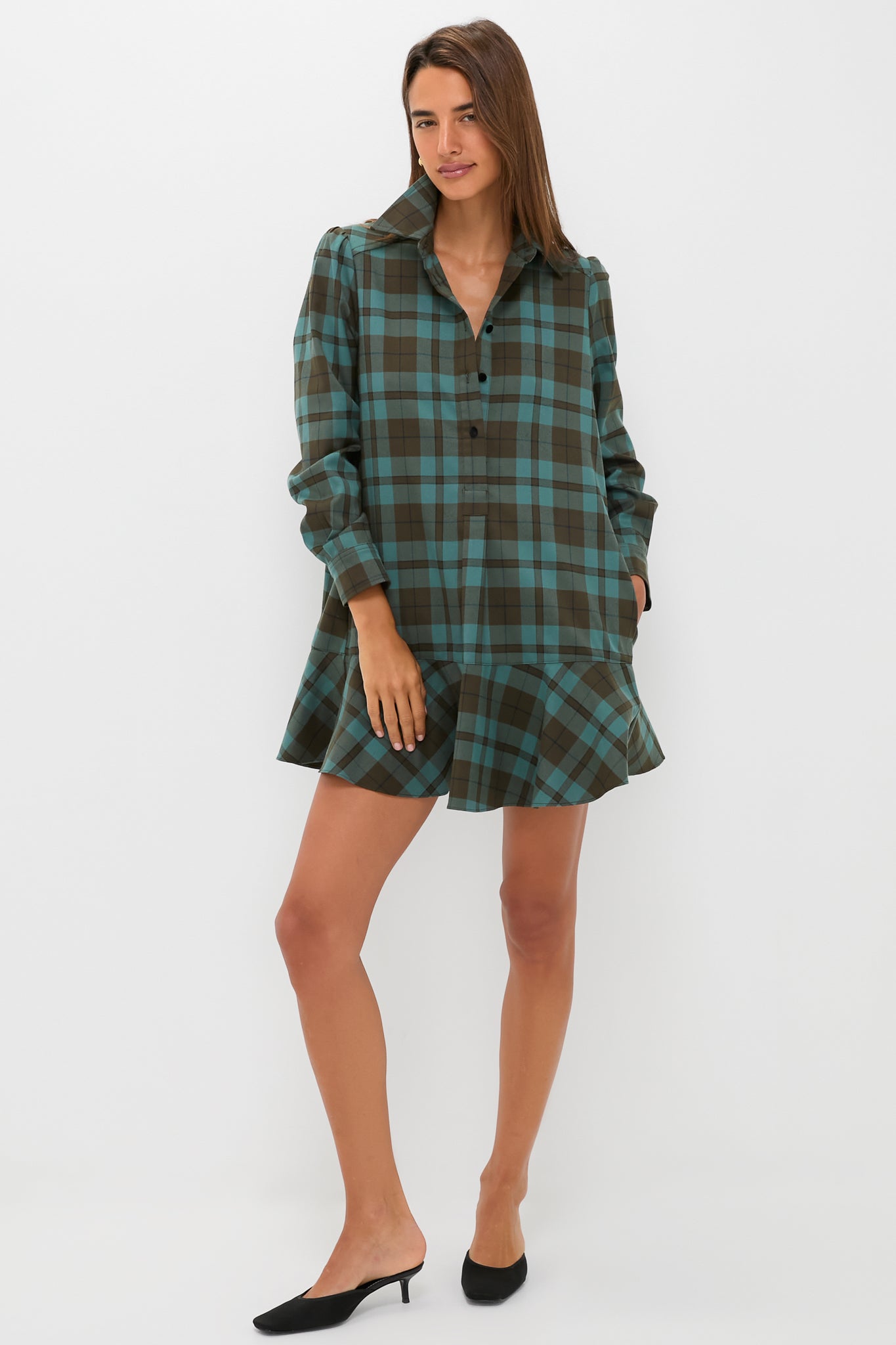 A woman poses against a white background wearing a Tuckernuck brown and teal plaid crepe shirt dress with a flared hem and relaxed fit, styled with black heeled mules.