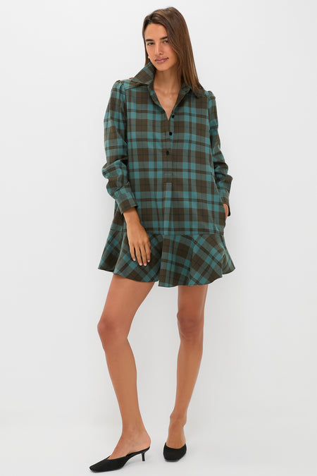 A woman poses against a white background wearing a Tuckernuck brown and teal plaid crepe shirt dress with a flared hem and relaxed fit, styled with black heeled mules.