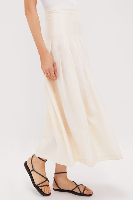 A model wears a light beige pleated maxi skirt, styled with a white top and black strappy sandals.