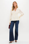 A woman stands against a white background wearing a Tuckernuck cream chiffon blouse with flared cuffs, long sleeves, dark blue wide-leg jeans, brown heels, and holding a small burgundy handbag.