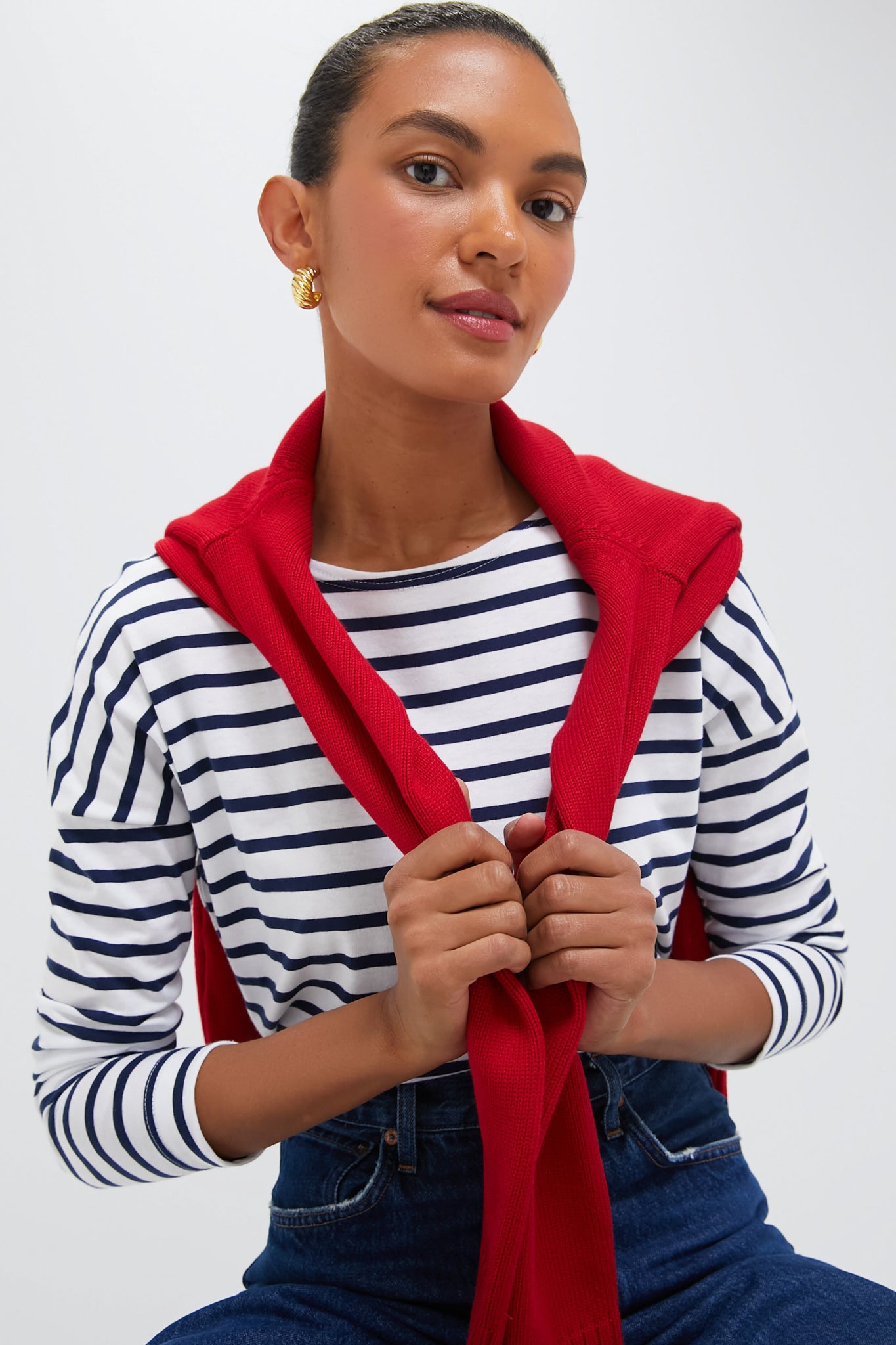 A woman wears a Saint James striped long-sleeve shirt, jeans, gold earrings, and has a red sweater over her shoulders as she looks at the camera.
