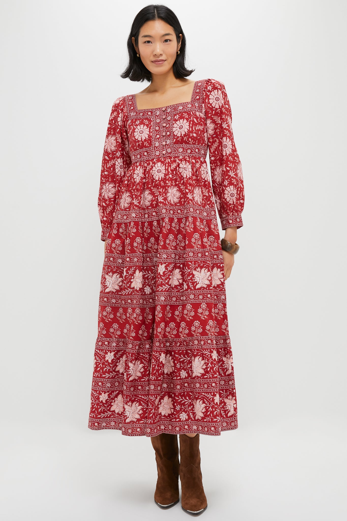 A woman wears a long red floral Lucia Dress by Pink City Prints, featuring long sleeves, 100% cotton fabric, and hand screen-printed details, paired with brown boots.