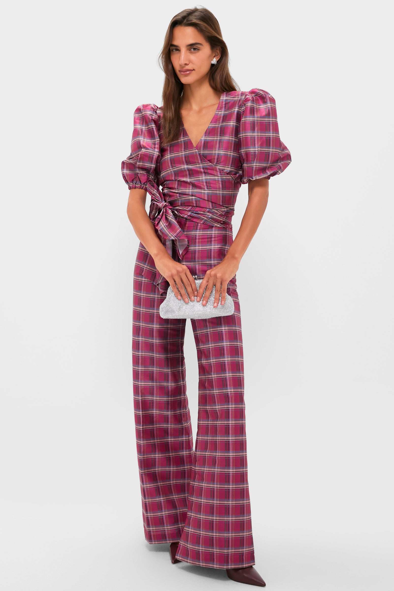 A woman wearing a pink and purple plaid wrap top with puff sleeves and matching flared pants holds an Olga Berg silver clutch, standing against a plain white background.