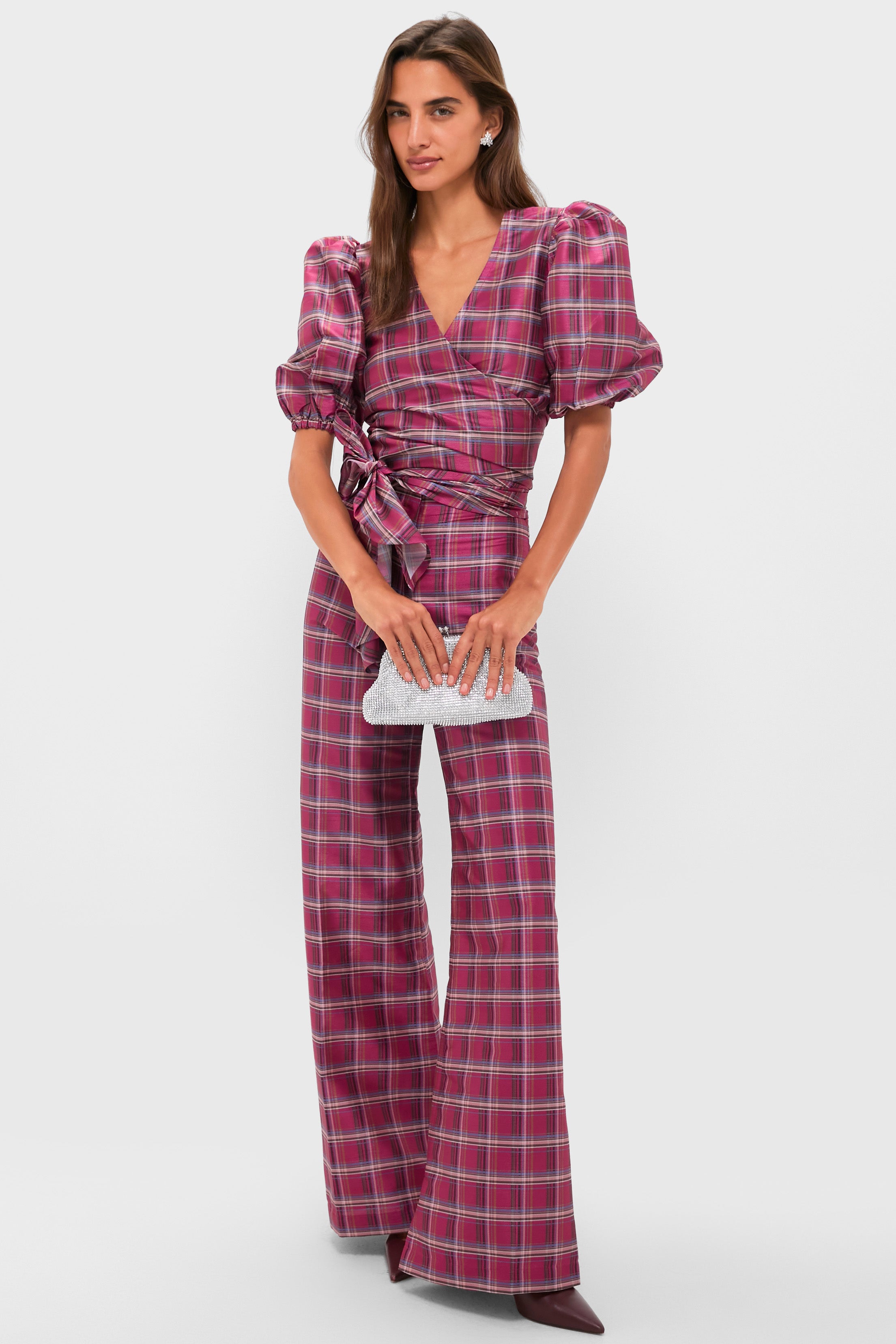 A woman wearing a pink and purple plaid wrap top with puff sleeves and matching flared pants holds an Olga Berg silver clutch, standing against a plain white background.