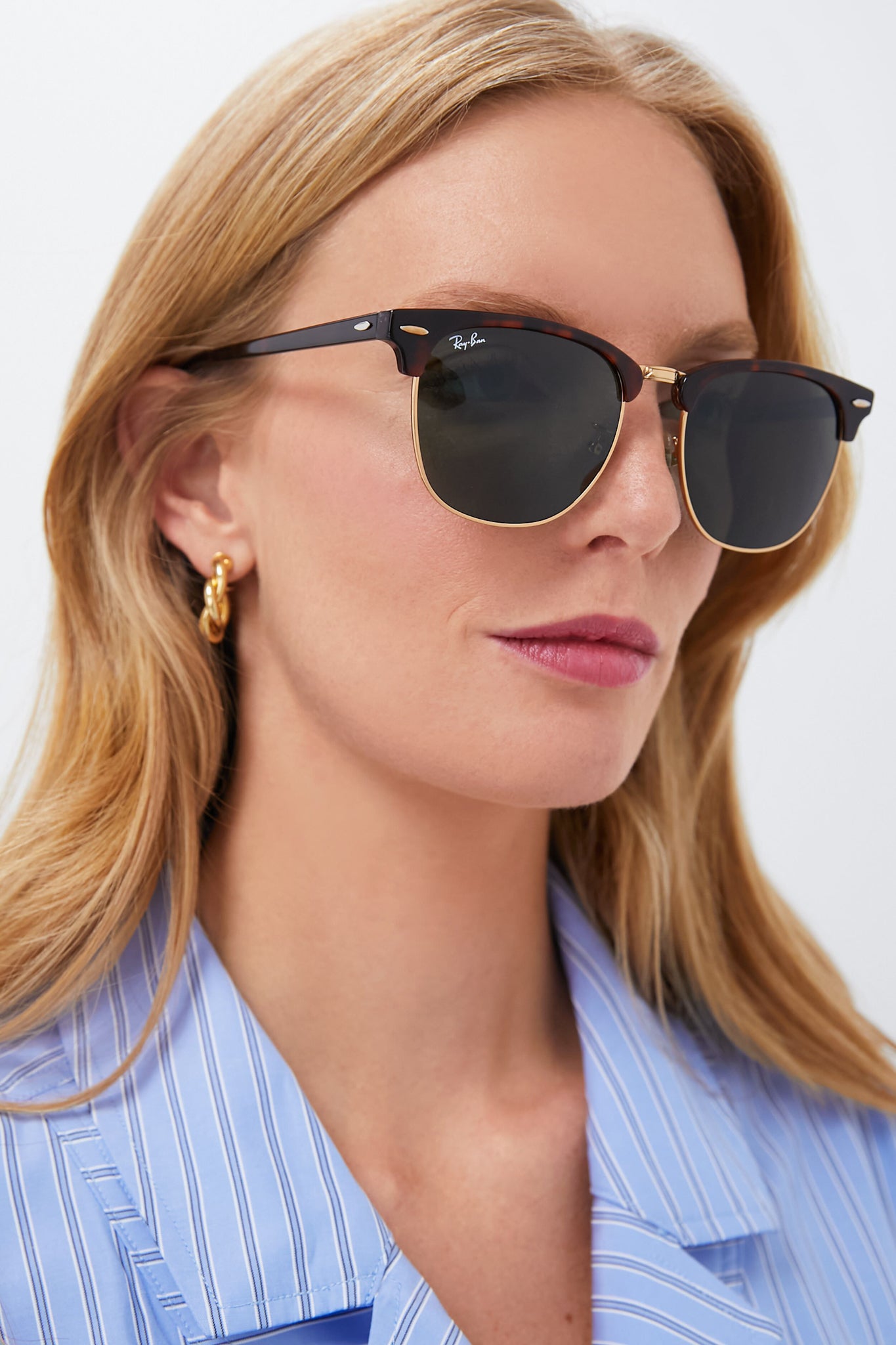 A blonde-haired woman wears Ray-Ban sunglasses with a timeless design, gold hoop earrings, and a blue pinstripe shirt against a plain background.