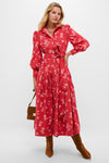 A woman models the Oliphant Nora Dress, a red floral long-sleeved cotton poplin midi with buttons and a tied waist, styled with brown knee-high boots and a handbag, posing against a plain backdrop.