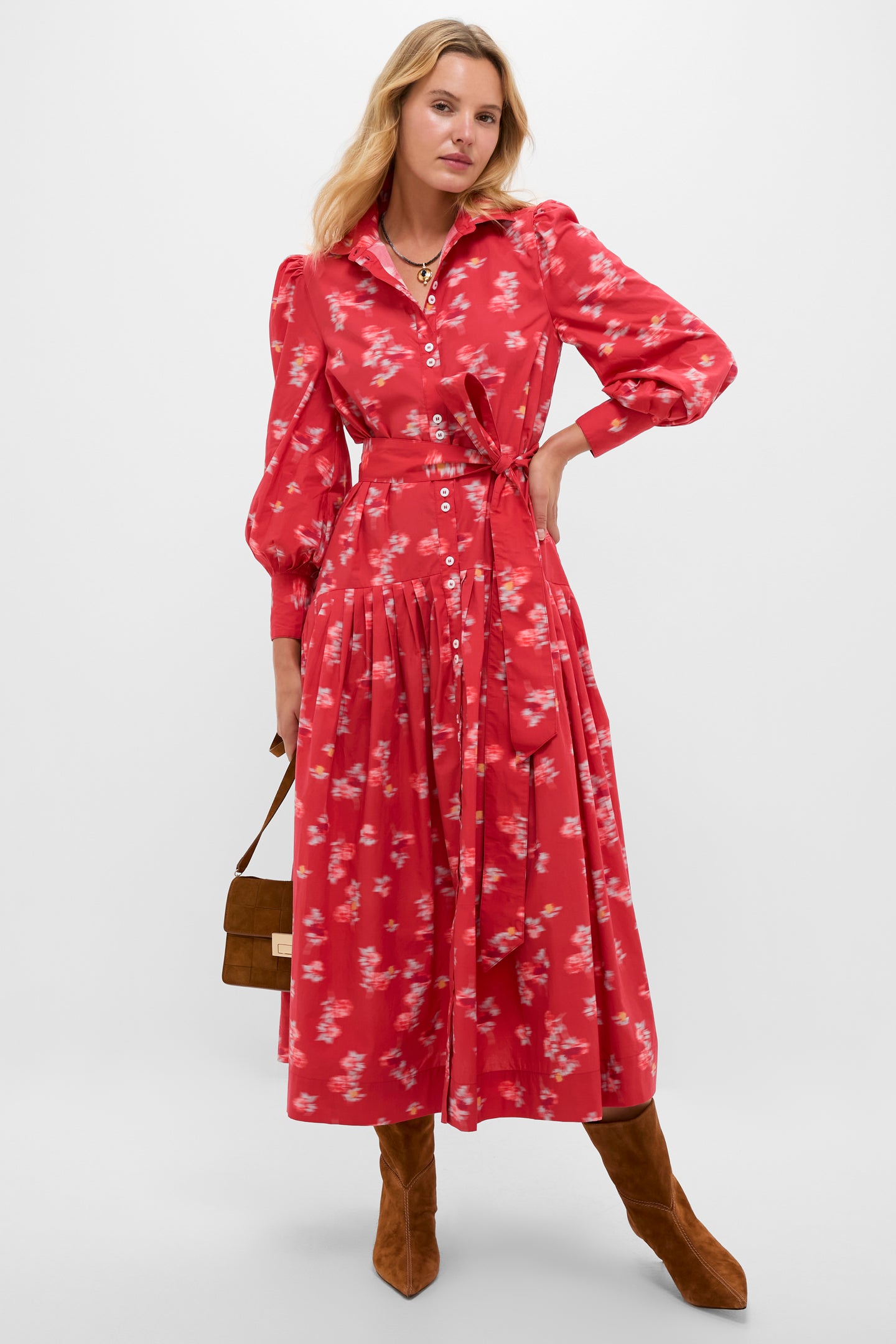 A woman models the Oliphant Nora Dress, a red floral long-sleeved cotton poplin midi with buttons and a tied waist, styled with brown knee-high boots and a handbag, posing against a plain backdrop.