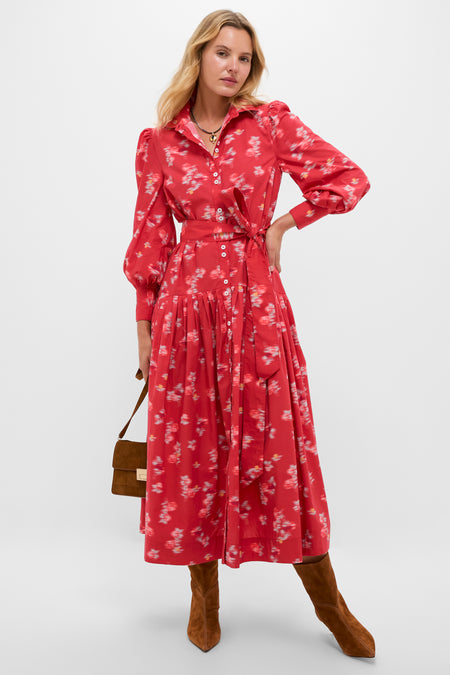 A woman models the Oliphant Nora Dress, a red floral long-sleeved cotton poplin midi with buttons and a tied waist, styled with brown knee-high boots and a handbag, posing against a plain backdrop.