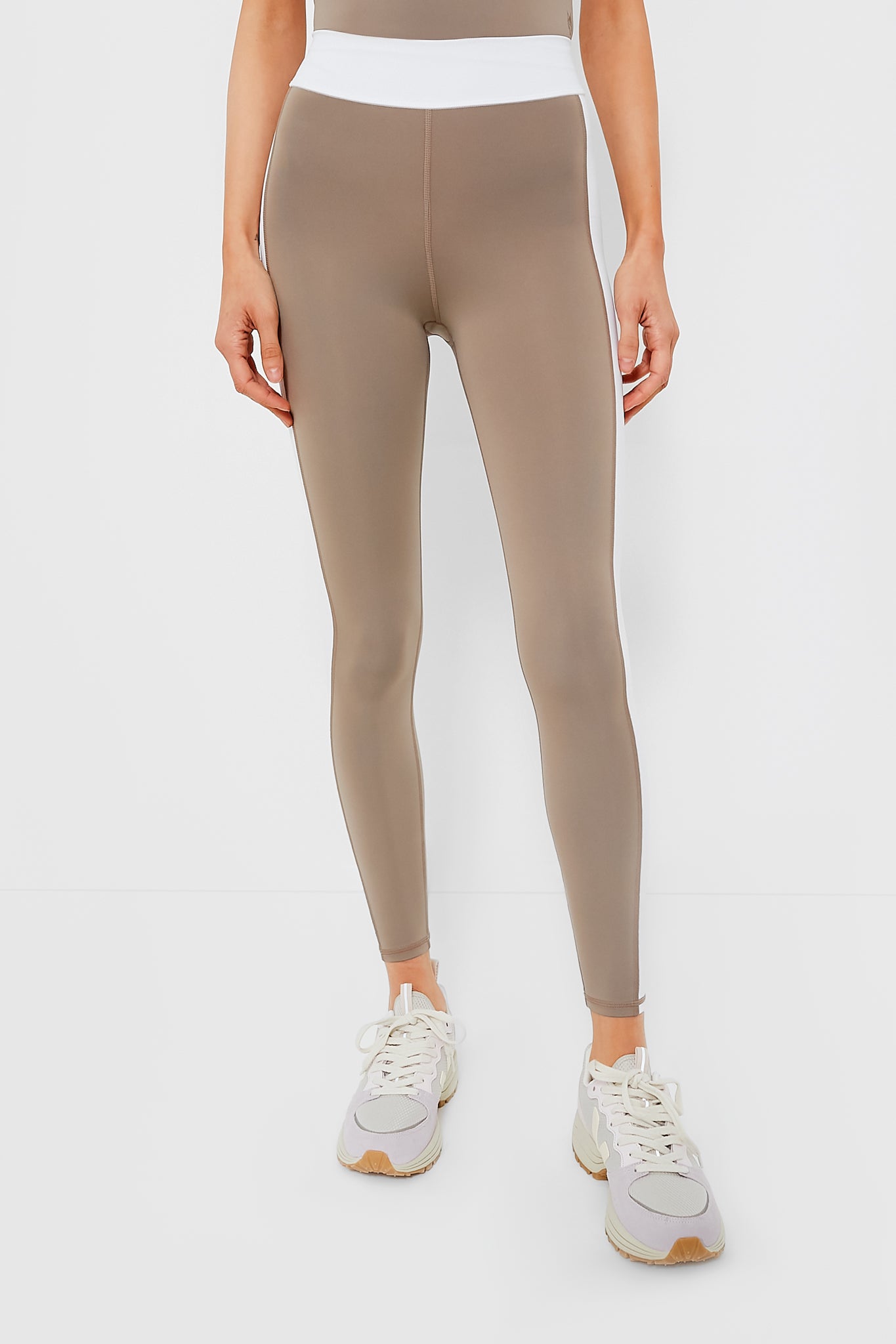 A person confidently wears Greyson Luna Legging high-waisted taupe leggings made from Italian fabric, featuring a white waistband and paired with white sneakers.