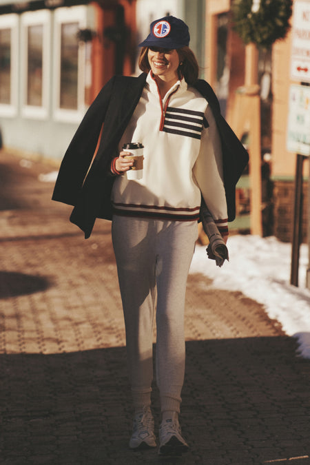 Young woman wearing the White Flag Heiden Quarter Zip with classic stripes and effortless style, enjoying a fun, sunlit street, embodying high-end, cool confidence.