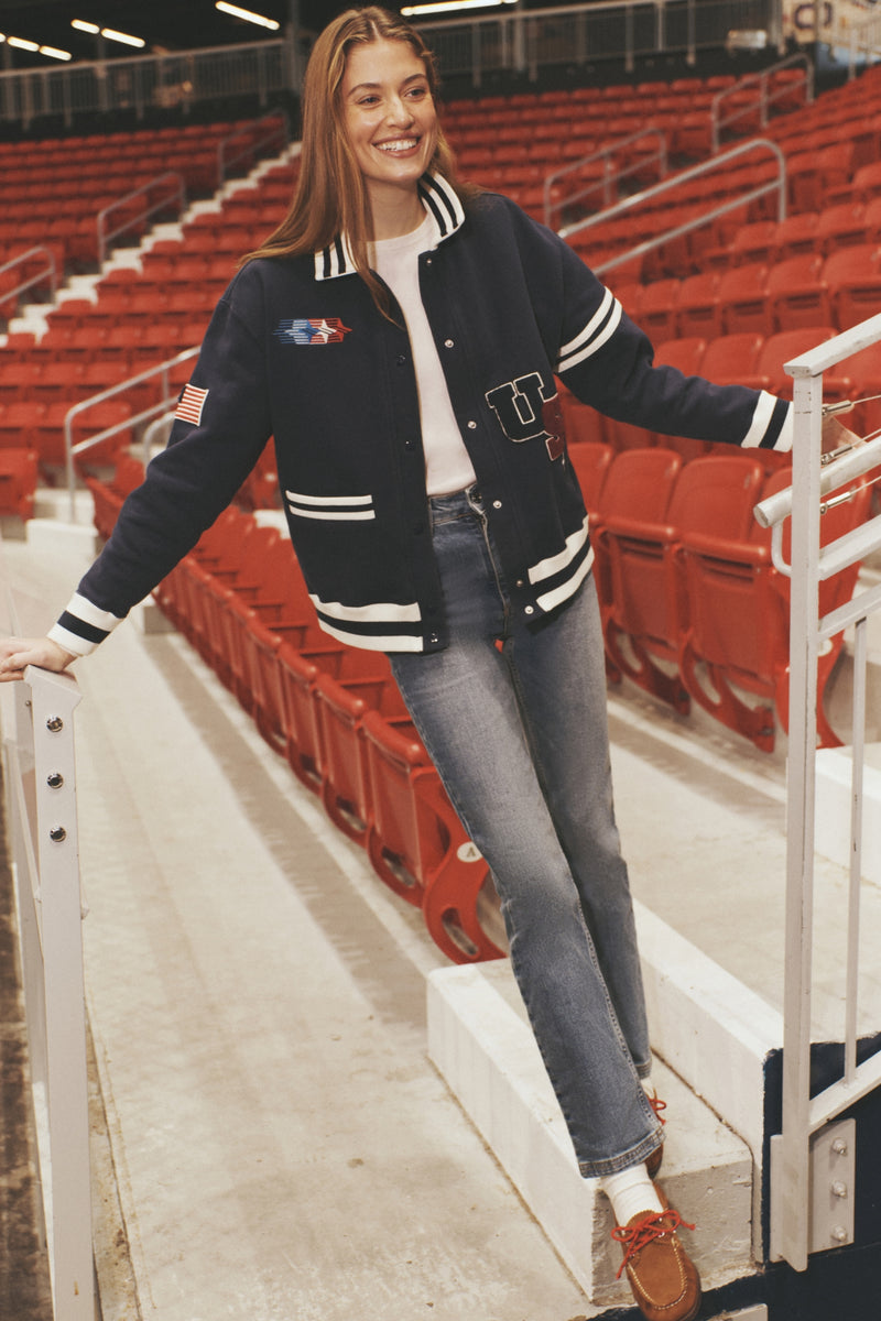 Young woman in a navy Albright Varsity Jacket with classic patches, embodying an effortless, fun, and high-end style in an empty stadium setting.