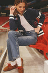 Young woman in a classic Navy Albright Varsity Jacket with patches, effortlessly adjusting an earring while seated in a fun, high-end stadium setting.
