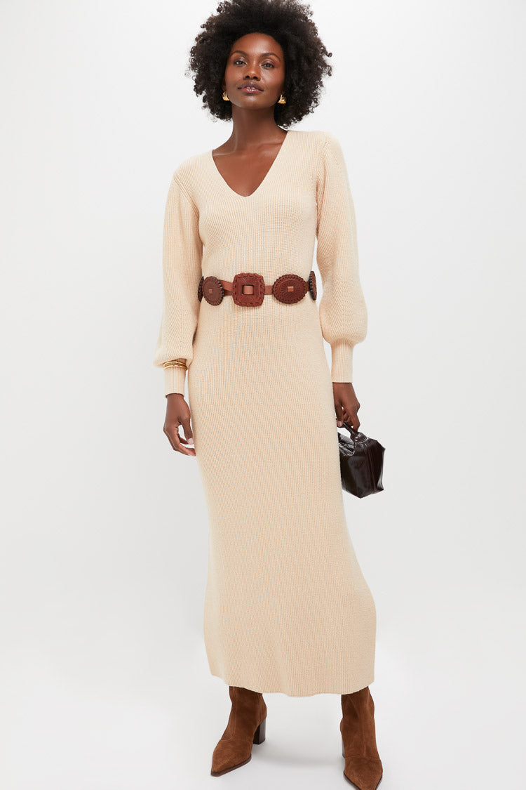 A woman in a beige Hyacinth House knit midi dress with a V-neckline, brown belt and boots, and carrying a dark handbag poses against a plain white background.