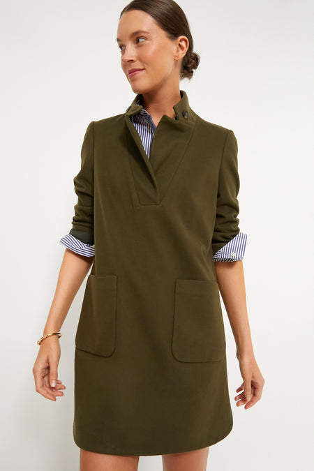 A woman wears the Tuckernuck Olive Emerson Dress—an olive green, long-sleeve dress with front pockets—over a blue striped shirt, channeling vintage equestrian style and serving as a timeless wardrobe staple.