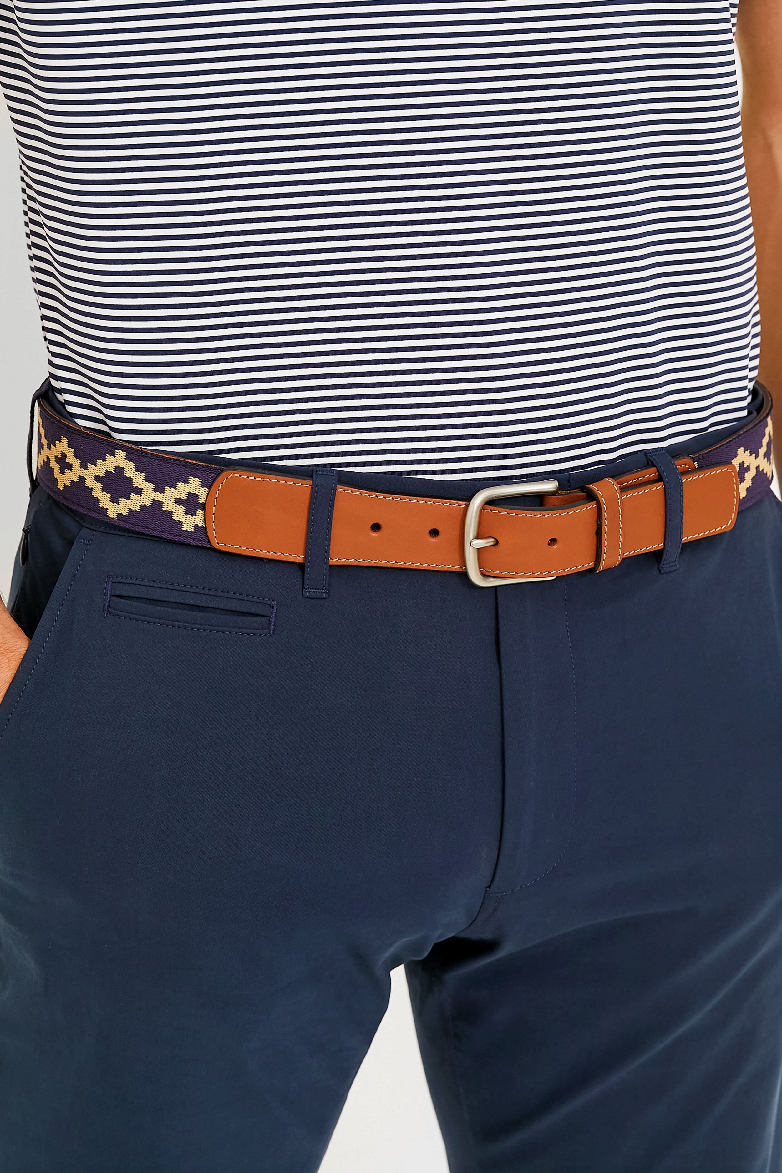 A person wears a navy and white striped shirt, navy pants, and a La Matera woven belt featuring Argentine fabric, tan vegetable-tanned leather accents, and a geometric pattern, with hands in pockets.