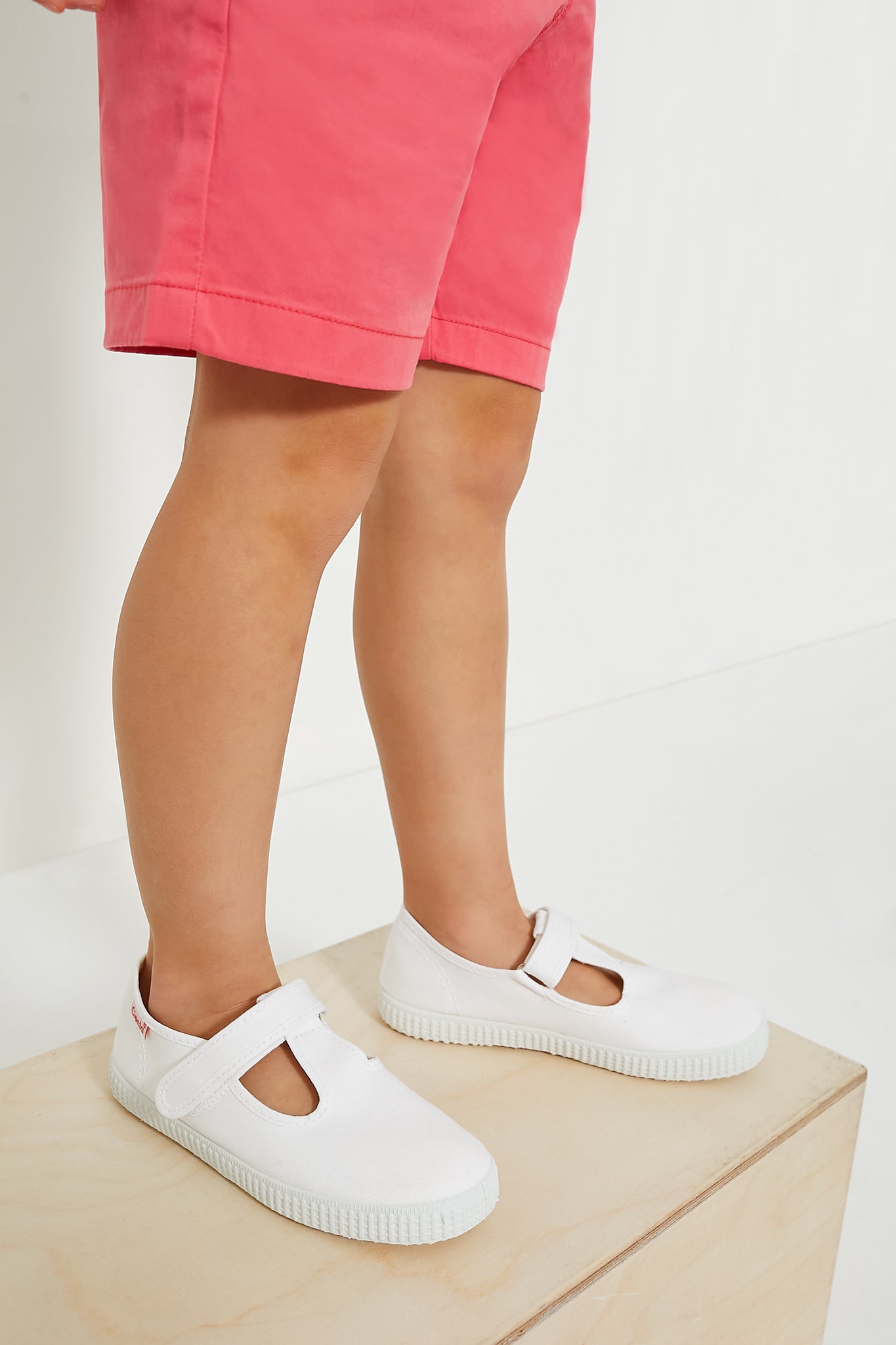A child in pink shorts and white Cienta vulcanized shoes stands on a light wooden block, highlighting stylish childrens footwear against a white background.