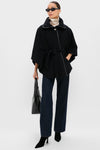 Wearing a ba&sh black belted wool cape coat, dark jeans, black heeled boots, sunglasses, and holding a black bag—an effortlessly chic nod to French fashion against a plain light background.